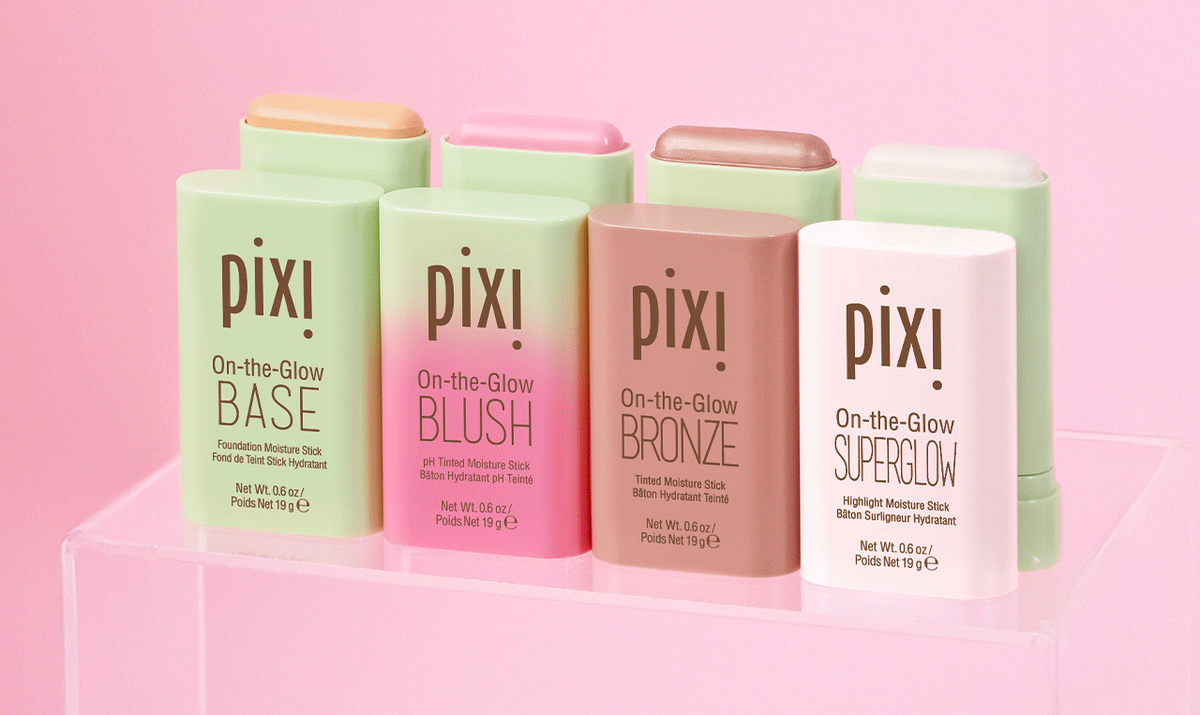 Four Pixi On-the-Glow foundation stick products arranged on a clear acrylic stand against a soft pink background. The products come in light green, pink-to-orange gradient, peach-to-pink gradient, and pale pink packaging, with the brand name 'pixi' clearly visible on each stick.
