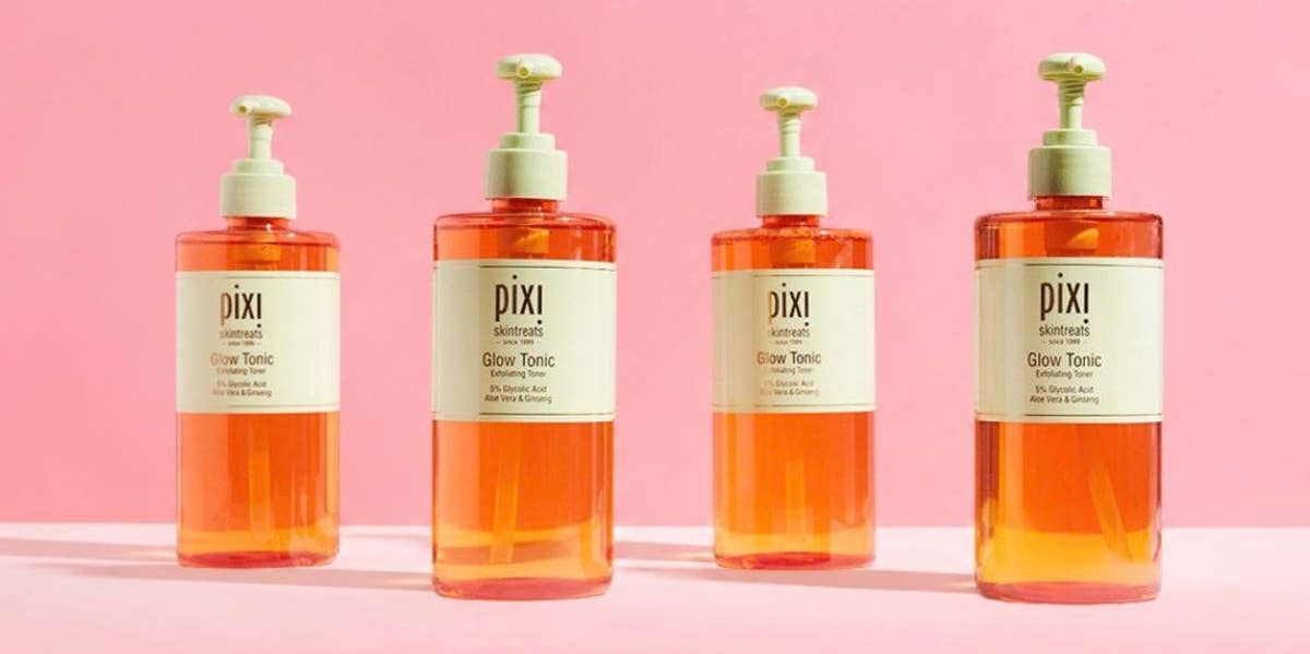 Four Pixi Glow Tonic pump bottles filled with orange liquid, arranged in a row against a solid pink background. View 2