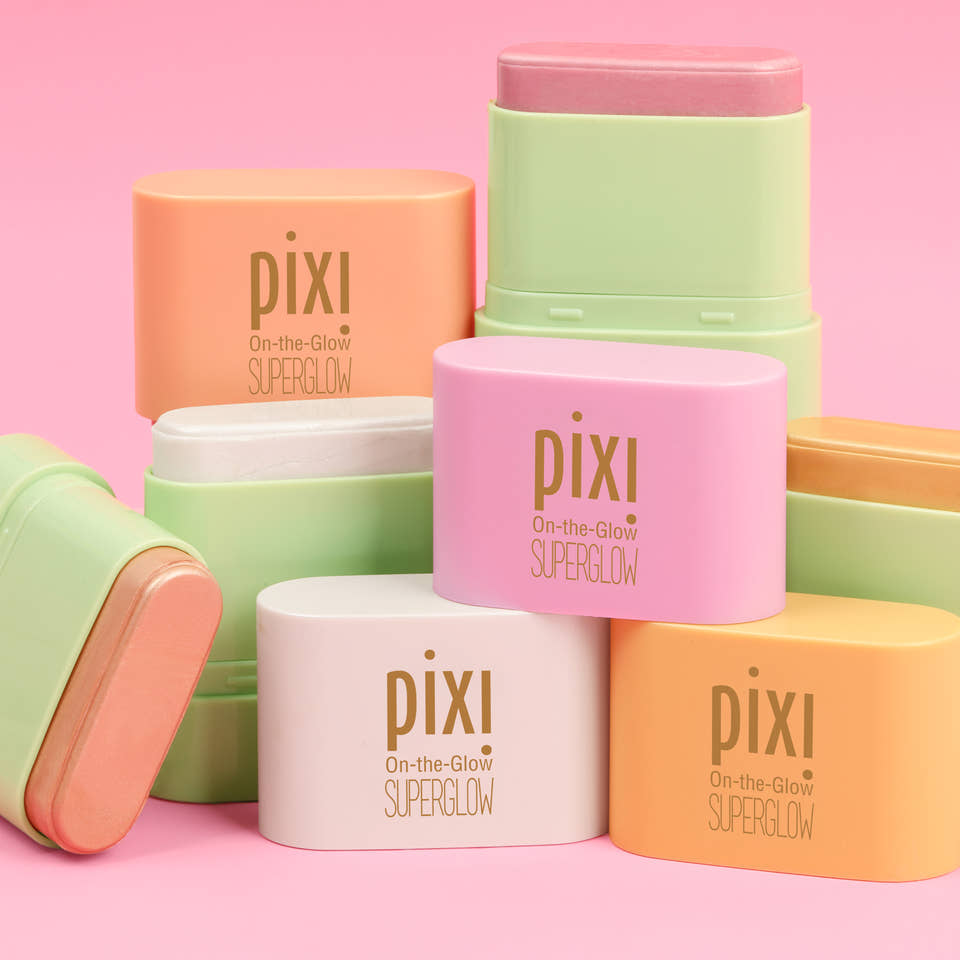 A group of Pixi On-the-Glow SuperGlow stick products in pastel shades of pink, orange, yellow, and white, arranged on a solid pink background - view 2 view 1