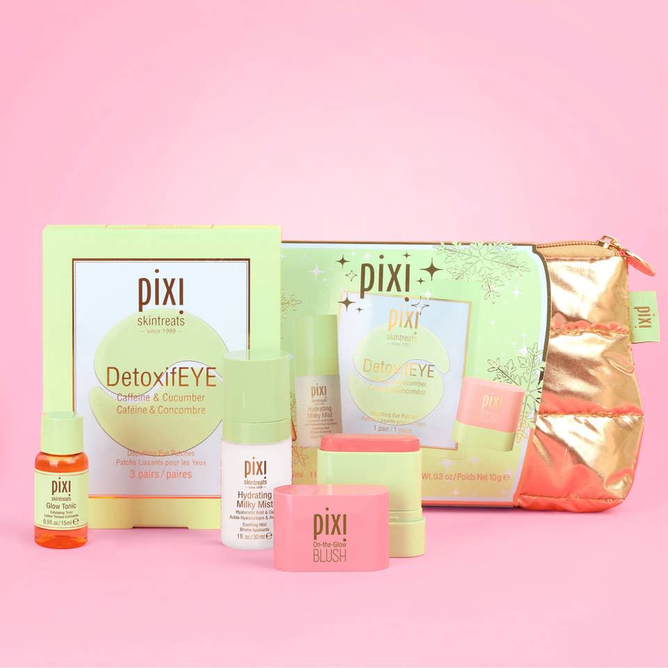 Pixi Skincare Heroes gift set arranged on a solid pink background, featuring a light green box, a metallic rose gold zip pouch, a small orange bottle, white and pink bottles, and a pink blush jar. view 1