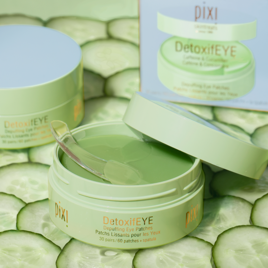 Pixi DetoxifEYE lifting eye patches shown in an open jar with a clear applicator spoon, alongside a closed jar, its packaging box, and sliced cucumber props on a light surface. view 1